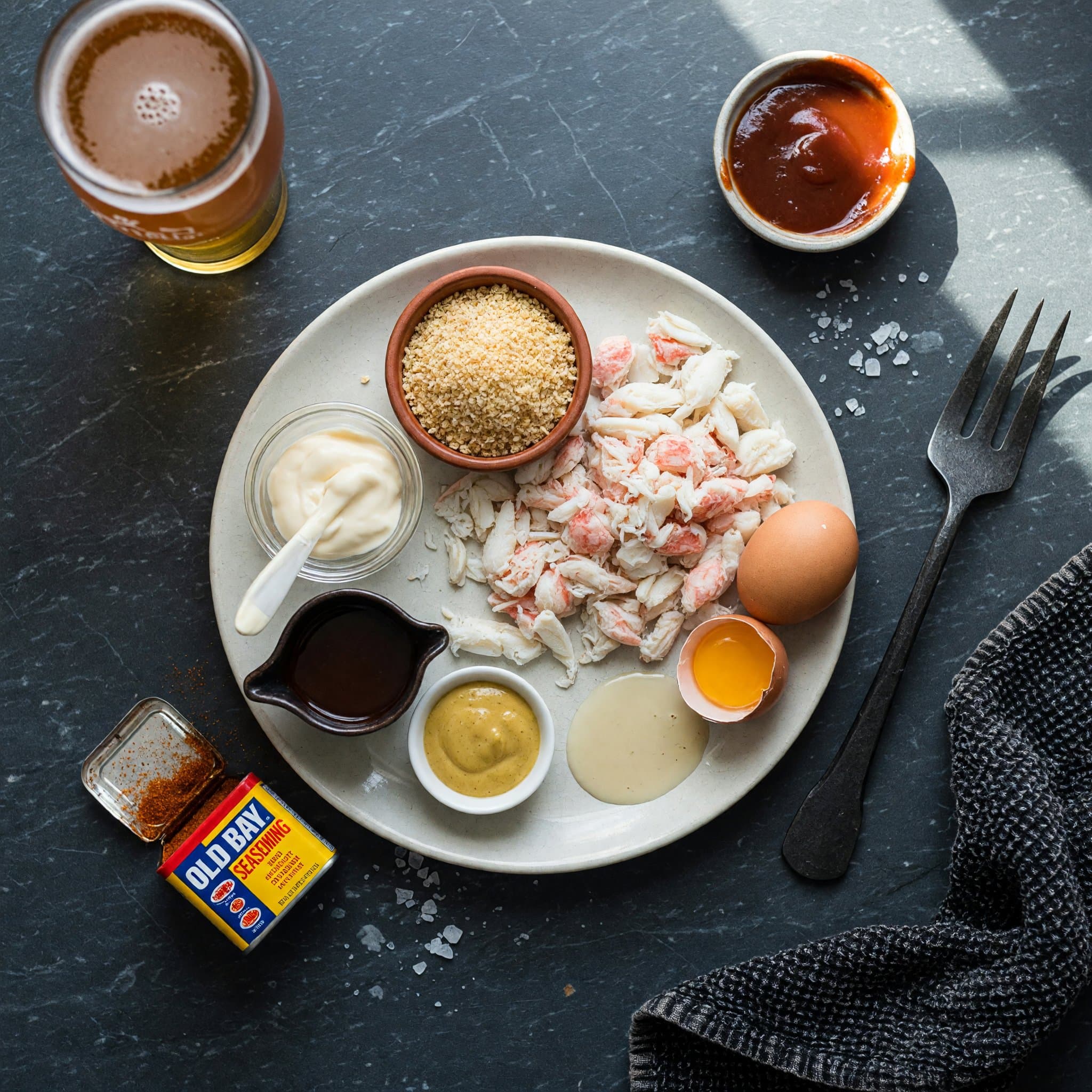 Overhead view of Lump Crab Meat, Breadcrumbs, Mayonnaise, Dijon Mustard, Worcestershire Sauce, Old Bay Seasoning, Egg and Salt arranged on a table